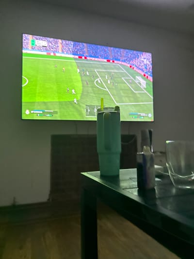 An indoor scene in Tristán Suárez, Argentina, displays a large screen projecting a soccer video game. The game shows a green pitch with white markings, a white goal, and multiple player avatars in white, dark, and yellow jerseys. Blue spectator stands are visible in the background of the virtual stadium. Game interface elements, including a "1-0" score and "37 CARRIAS" player information, are visible on the screen. In the lower right foreground, a dark rectangular table holds a large, light blue or teal insulated cup with a yellow straw, a small dark object resembling a lighter or bottle, and a partially visible clear glass. The room features light-colored walls, a dark, textured surface (possibly brickwork) behind the table, and a light-colored floor. The primary illumination originates from the screen.
