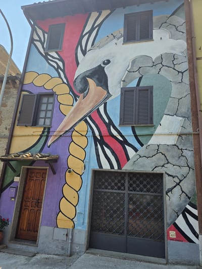 The image depicts the exterior of a building in Viterbo, Italy, adorned with a vibrant mural. The mural features a stylized, large white bird's head with a prominent beak, rendered in shades of white, grey, and black. The bird's form is integrated into a dynamic composition of colorful geometric shapes and abstract lines in red, blue, yellow, purple, and green. A thick, yellow, braided rope element is prominent on the left side of the mural. The building has two ground-level entrances: a wooden door on the left, set within a purple-painted recessed area, and a dark metal gate on the right. Above these entrances, there are two windows with dark shutters on the left and one on the right. The building's facade is textured, and the mural art incorporates elements that mimic cracks in stone. A signature "TINA LOIODICE" is visible at the bottom right of the mural. The surrounding environment shows a paved walkway and parts of adjacent buildings.