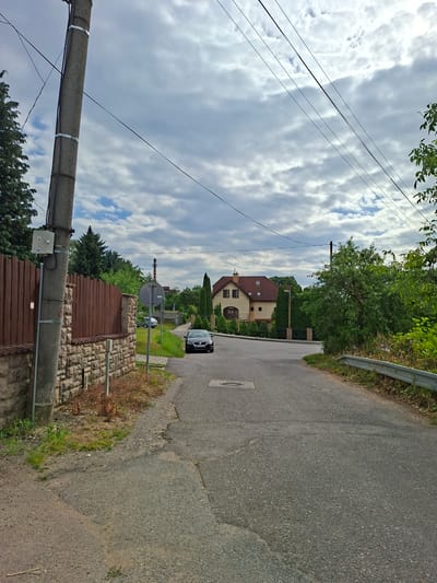 The image depicts a quiet, tree-lined street in Ilava, Slovakia, on a cloudy day. A dark-colored sedan is parked on the right side of the road, facing away from the camera. To the left, a concrete utility pole stands prominently, adorned with electrical boxes and wires stretching across the frame. Adjacent to the pole is a stone wall topped with a wooden fence. In the background, a two-story house with a red tile roof is visible, surrounded by manicured hedges and trees. The road itself appears to be made of asphalt and shows signs of wear and tear. The overall atmosphere is one of suburban tranquility.