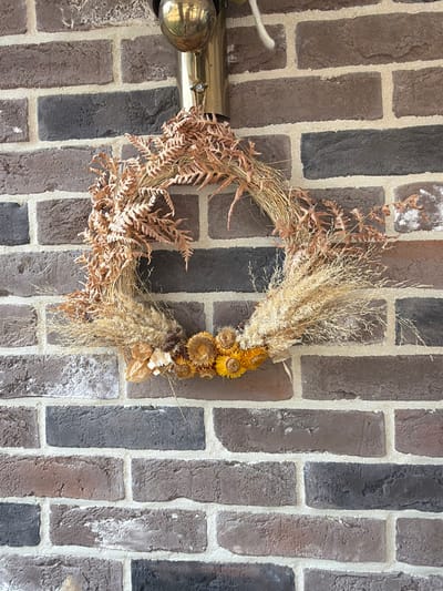 A rustic wreath made of dried ferns, grasses, and dried flowers is hanging on a brick wall. The wreath has a circular base with various dried botanical elements arranged around it. The brick wall is made of dark brown and grey bricks with light colored mortar. Above the wreath, a brass-colored lamp fixture is visible. The image appears to be taken outdoors, possibly on a porch or exterior wall. There are no people or animals present in the image.