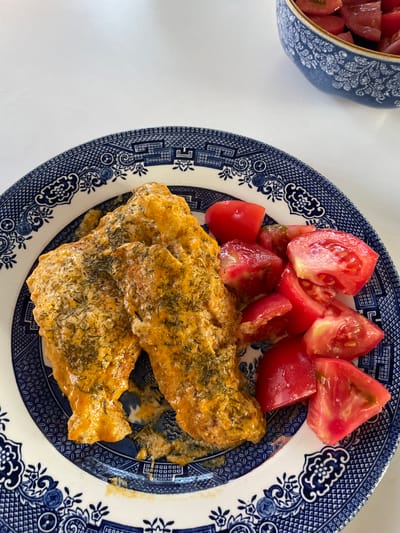 The image shows a close-up, overhead view of a blue and white patterned plate holding two pieces of food coated in a creamy, orange sauce and sprinkled with dill. To the right of the food are several chopped red tomatoes. In the background, a blue and white patterned bowl filled with more chopped tomatoes is visible. The setting appears to be a white table. The location is not specified as Skala Avdiron, Greece, but is a domestic indoor setting.