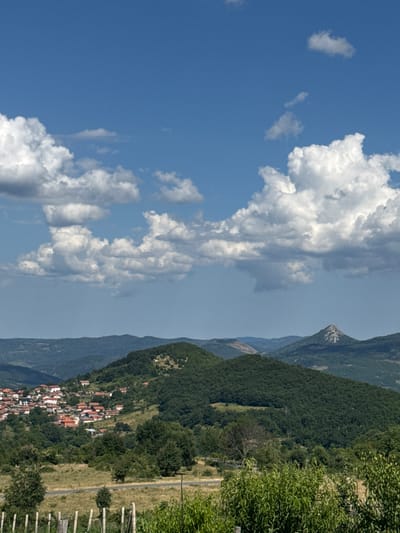 This is a vertical, medium-shot, outdoor photograph of a village nestled on a hillside in Karaveliovo, Bulgaria. The sky is a bright blue with scattered white, fluffy clouds. In the mid-ground, a village with red-tiled roofs and white-walled houses is spread across a green, tree-covered hillside. A paved road winds through the scene. In the background, rolling, forested hills recede into the distance, with a distinct, rocky peak visible on the far right. The foreground features a wooden fence and lush green foliage. The scene is illuminated by natural daylight, suggesting a sunny day.