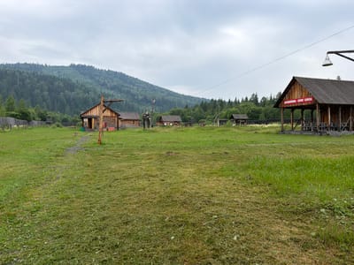 The image depicts an outdoor scene in a rural setting, likely a village or an agricultural compound, under an overcast sky. In the foreground, a large expanse of mown grass covers the ground, with patches of longer grass and some scattered wildflowers on the right. A narrow gravel path winds through the grass towards a cluster of wooden buildings.

To the left, a rustic wooden fence encloses an area, and a small wooden guard tower or observation post stands within it. Several log cabins and smaller wooden structures are visible, appearing to be part of a settlement. One of these buildings has a sign with Russian text in red.

In the midground, more wooden structures are scattered across the grassy field. A prominent wooden building with a large open-fronted section, possibly a covered market or workshop, is situated on the right side of the image. A large, cone-shaped lamp is suspended from a metal pole above this structure.

The background is dominated by a dense, green, tree-covered mountain range, indicative of a forested, mountainous region. The overall impression is of a remote, possibly historical or traditional settlement, nestled within a natural landscape. The location is identified as Ust'-Kabyrza, Russia.