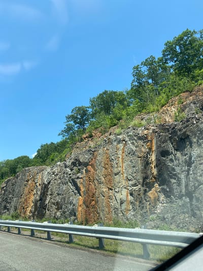 The image shows a roadside rock face in Pointe-au-Chêne, Canada. The rock face is steep and jagged, with streaks of orange and brown iron staining. Trees and vegetation grow along the top and sides of the rock face. A metal guardrail separates the road from the rock face. The road appears to be paved and is visible in the foreground. The sky is blue with a few wispy clouds. The overall impression is of a natural landscape that has been modified for road construction.