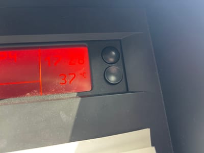 The image shows a close-up view of a car's dashboard. The main subjects are two round, black buttons and a red digital display. The display shows "37 C" on the top line and "17:28" followed by what appears to be "P4" on the bottom line. The context is a car's interior, likely in Espalion, France, though no external location cues are visible. The display indicates a time of 17:28 and a temperature of 37 degrees Celsius. The image is a still photograph with no discernible activity or event taking place beyond the passive display of information. Notable details include the dust on the dashboard and the display screen, suggesting the car may not be brand new. The lighting suggests it is daytime, but the exact time of day is further clarified by the display. No people are visible in the image, and no emotions can be inferred. The visible text on the display is "37 C" and "17:28 P4".