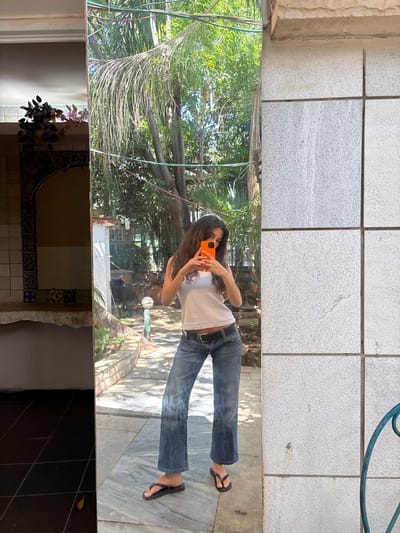 A young woman is standing in front of a full-length mirror, taking a selfie with her smartphone. She is wearing a white tank top and blue jeans. The woman is facing the mirror, with her back to the viewer. She is standing on a tiled floor. There is greenery visible in the mirror's reflection. The location is in the city of Ra'anana, Israel, with the coordinates 32.173, 34.872.  The image is taken in what appears to be a courtyard or patio area.  The wall behind the mirror is made of white tiles.  There is a blue metal object on the wall to the right of the mirror.  The woman is smiling in the selfie.