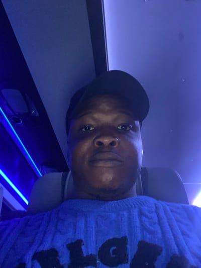 A person with a cap sits in a dimly lit space with blue lighting. They appear to be in a car or vehicle with a built-in entertainment system. A patterned blue sweater or blanket is draped over the individual. The lighting and composition give the impression of a relaxed or possibly sleeping posture. The image is taken from a low angle, slightly tilted.