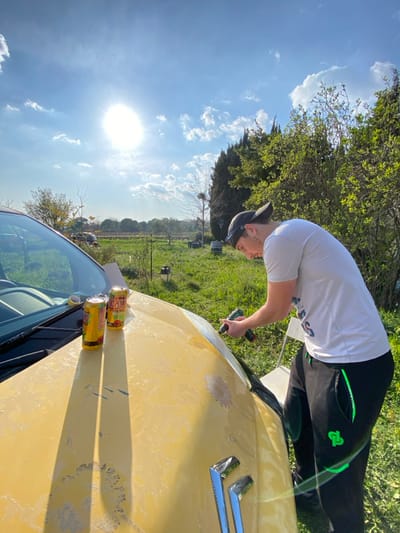 The scene is an outdoor environment in Cucuron, France, illuminated by direct sunlight under a blue sky with scattered white clouds. A male subject, dressed in a light grey short-sleeved shirt, dark track pants with green detailing, and a dark baseball cap worn backward, is depicted leaning over the front of a light yellow vehicle. He is holding a green and black handheld power tool, applying it to the vehicle's hood, which exhibits areas of chipped and peeling paint, revealing underlying material. Two yellow and red aluminum beverage cans, labeled "Damm Lemon" and "Damm Milano", are resting on the vehicle's hood, casting distinct shadows to the left. A roll of yellow adhesive tape is visible near the base of the windshield. The background features a grassy field containing various trees and shrubbery, including a large nest in a leafless tree. A white plastic chair is partially visible behind the male subject.