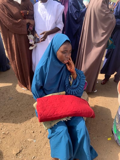 The image shows a gathering of people in what appears to be an outdoor location in Naraguta Hausawa, Nigeria. Predominantly women are visible, dressed in traditional Islamic attire, including headscarves (hijabs) and gowns in varying colors such as blue, brown, and white. One woman is lying down, resting her head on a red fabric. Her hand has henna designs on it. Other people are standing around her. The ground is dirt or gravel. Some accessories such as keychains are visible in people’s hands.