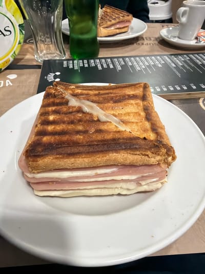 The scene depicts an indoor dining setting at Panda Café & Bar in Gondomar, Portugal, identified by "PANDA" branding on a menu. In the foreground, a large, rectangular toasted sandwich (tosta mista/especial) with prominent grill marks rests on a white, oval-shaped plate. The sandwich has a golden-brown crust; a diagonal cut on the top reveals melted white cheese oozing out. Multiple distinct layers of light pink ham and white cheese are visible between slices of toasted bread. Small crumbs are scattered on the plate. To the left, a clear, ribbed drinking glass and a partially visible dark green bottle are present. To the right, a dark menu board with white text and numeric prices, indicating costs such as "7.00€" and "11.50€," lies flat on a light brown table surface. Further back, a second, partially visible toasted sandwich on a smaller white plate, a white ceramic coffee cup on a saucer, and a small, crumpled orange and white foil packet are arranged. A paper placemat with "SUPER ES" branding is also visible. No individuals are clearly depicted, but the arrangement of food and drink suggests active dining.