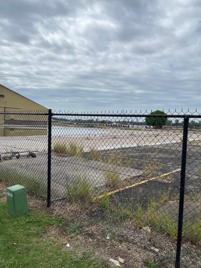 This image, taken in Hamilton, Australia, presents an outdoor scene viewed through a prominent black chain-link fence running horizontally across the frame. The scene is captured on an overcast day, with the sky filled with textured grey clouds.

In the foreground, to the right of the fence, there is an area of green grass with some patches of dry vegetation and scattered litter. A rectangular green utility box with an unreadable white label sits on this grass in the bottom-right corner. Near the bottom-center, some metal pipes or conduits are visible on the ground directly in front of the fence.

Beyond the chain-link fence, a large paved or asphalt area stretches into the distance. This surface appears worn, cracked, and discolored, suggesting disuse or neglect. Faded yellow lines are intermittently visible on the pavement. Sparse, wild grass and weeds grow through cracks in the ground and along the edges of the paved area. A small, dense, rounded green tree or bush stands behind the fence towards the left-center of the image. The corner of a light-colored building with a sloped roof is visible in the bottom-left, behind the fence and pipes. The distant horizon appears hazy under the cloudy sky. No people are present in the scene, and the overall atmosphere suggests a functional yet somewhat neglected or abandoned industrial-like boundary area.