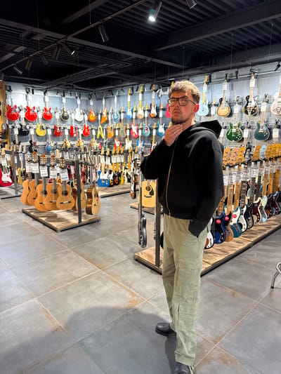A fair-skinned man with short, curly blond hair and glasses stands in a guitar store. He wears a black hoodie and light green cargo pants, with black shoes. He looks toward the camera and has his left hand near his face.

The background consists of displays of guitars on both the wall and on racks, showcasing various colors and styles. The store has a modern aesthetic with a high ceiling and industrial-style lighting fixtures. The floor is tiled with large, gray tiles.

The location is a retail space likely in Palaiseau, France.