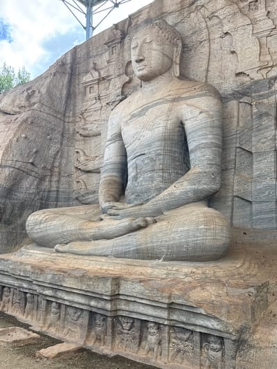 The image shows a large stone statue of a seated Buddha, located in Polonnaruwa, Sri Lanka. The statue, carved directly into a rock face, depicts Buddha in a meditative pose, with legs crossed and hands resting in the lap. The stone has a striated texture, with varying shades of gray. The environment surrounding the statue is also rock, and some greenery can be seen to the lower left. Above the statue is a metal support structure and blue sky.