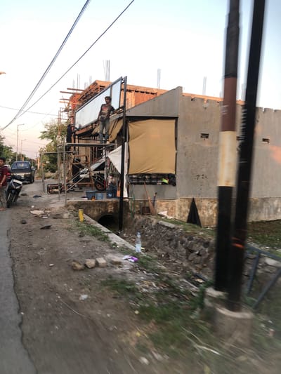 The image depicts a construction site in Sentul, Indonesia. In the foreground, a paved road runs alongside a dirt and gravel area with scattered debris. A motorcycle is parked on the left side of the road. Further into the scene, scaffolding is erected around a partially constructed building. Two men are visible on the scaffolding, one appearing to be working on a large sign or billboard. Another man stands on the ground near the scaffolding. The building itself is a concrete structure with exposed brickwork and openings. A ditch runs along the side of the construction area, and a large black pole with a white stripe is prominently positioned in the right foreground. The sky above is clear and shows the warm hues of a sunset.
