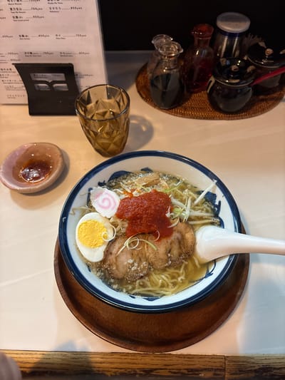 A bowl of ramen sits on a wooden tray on a white table. The ramen contains noodles, broth, pork slices, green onions, a pink fishcake, a hard-boiled egg, and a dollop of red chili paste. A white spoon is placed diagonally across the bowl, partially submerged in the broth.  A glass of water sits to the left of the bowl, with a small dish of red sauce in front of it.  Four condiment bottles, two dark brown, one red, and one silver, are visible on a woven placemat in the upper right corner of the image. The table is made of light wood with a grain pattern.  The location of the image is Chūō, Japan, specifically at the coordinates 35.687, 139.785.  The scene is a typical Japanese restaurant table where someone is eating ramen.