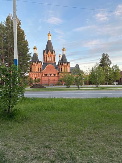 The image shows the Church of the Icon of the Mother of God of Kazan, located in Uray, Russia. The red brick church is on the left side of the frame, featuring multiple golden domes. A two-lane road separates the church from a green grassy area and sparse trees on the right. The sky is visible above the church, featuring a mix of blue and white clouds. A few trees are in the foreground at the bottom, along with a white and blue pole.