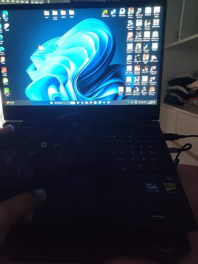 This image depicts an indoor scene within the city of Partido de Quilmes, Argentina. The primary subject is a dark-colored laptop, open and displaying the Windows 11 desktop with a blue abstract wallpaper. Numerous application icons, predominantly for gaming platforms and titles (e.g., Steam, Epic Games, Resident Evil 4, Red Dead Redemption 2, Elden Ring, Hogwarts Legacy), are visible across the screen. The taskbar shows the time "10:58" and the date "1/3/202x," with "Buscar" (Spanish for "Search") in the search bar. Stickers on the laptop's palm rest indicate "Intel Core" and "NVIDIA GeForce RTX" processing capabilities, and a faint "ROG" logo is visible on the screen bezel. A human hand, light-skinned, is visible in the lower-left portion of the frame, holding a black gamepad. The gamepad is connected to the laptop via a cable plugged into the right side. Another cable is also plugged into the laptop's right side. In the dimly lit background to the right, a piece of white or light-colored furniture, possibly a chest of drawers or shelf, is visible, with several folded items of clothing or fabric stacked on its surface. The overall environment suggests a personal living space, likely a bedroom or study.