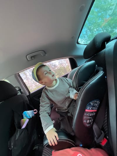 The image shows a young child in a car seat in the back seat of a car. The child is wearing a striped shirt and a yellow and blue hat, and is looking upwards. There are two car seats visible in the back of the car. A black bag with some items in it, including what appears to be a pack of wipes, is hanging on the side of one of the car seats. The car appears to be moving as the trees and sky visible through the windows are blurred. The location is identified as Bolgury, Russia.