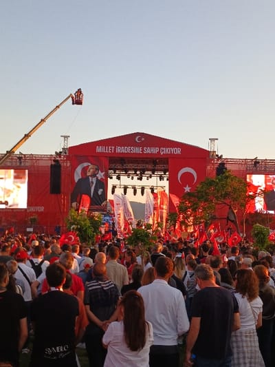A large crowd of people gathered in front of a stage with a large red banner that reads "Millet İradesine Sahip Çıkıyor" (The Nation Stands Up to Its Will). The stage is adorned with Turkish flags and the image of a man in a suit, likely a political figure. The scene takes place in the Konak district of İzmir, Türkiye. The crowd is dense and comprised of individuals of all ages, dressed in casual clothing. Many are holding Turkish flags, some are taking pictures or videos of the stage and the crowd. There is a large crane to the left of the stage with two individuals working on the crane. The scene is likely a political rally or public event, held outdoors under a clear sky.
