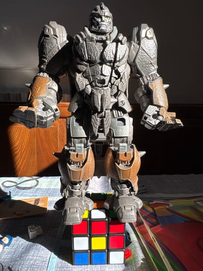This is a photograph of a large, detailed toy robot figure placed next to a Rubik's Cube. The robot appears to be made of plastic and metal, with intricate mechanical details and a weathered, metallic finish. It has a complex design with multiple articulated limbs and a segmented body. The Rubik's Cube is partially solved, showing a mix of colored squares: red, yellow, blue, and white.

The scene is lit by a strong light source coming from the left, casting long shadows, suggesting it is either morning or late afternoon. The background is dark, possibly a wall, with a wooden door visible to the right, indicated by the warm tones and grain pattern. A colorful patterned fabric, possibly a blanket or tablecloth with abstract designs and what appears to be a partial illustration of a person in superhero attire, covers the surface where the robot and cube are placed. There are also small toy building blocks and a miniature wooden door prop visible.

The photograph is taken from a top-down perspective, with the robot figure oriented horizontally across the frame. The overall impression is of a still-life arrangement, possibly a toy display or a child's play area. No people are visible, but the presence of toys suggests a domestic or play setting. The specific location cues like the wooden door and the style of the patterned fabric do not strongly suggest the provided location context of Sant'Angelo di Piove di Sacco, Italy.