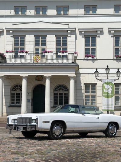 The image shows a white convertible Cadillac Eldorado parked on a cobblestone street in front of a white, neoclassical building. The building has multiple windows, a balcony, and columns. A banner with the text "Peace Mayors for Peace" and "Bürgermeisterinnen und Bürgermeister für den Frieden" is hanging from a lamppost. A coat of arms is visible above the entrance. The car's license plate reads "HL E 72 H". The scene appears to be in the city of Wismar, Germany, specifically at the Wismar City Hall (Rathaus). There are no people visible in the image. The time of day appears to be daytime, with bright sunlight casting shadows.