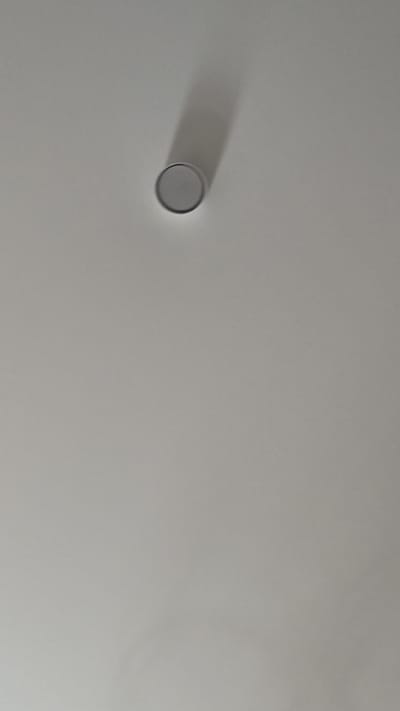 A single, circular object with a metallic sheen is affixed to a plain, light-colored surface, likely a ceiling. The object is casting a long, elongated shadow, indicating a light source from the upper left. The surface appears smooth and is uniformly lit, with no other discernible features or objects present in the frame. The image does not provide any contextual information about the location within the city of Kovrov, Russia, or any details related to people, actions, or interactions.
