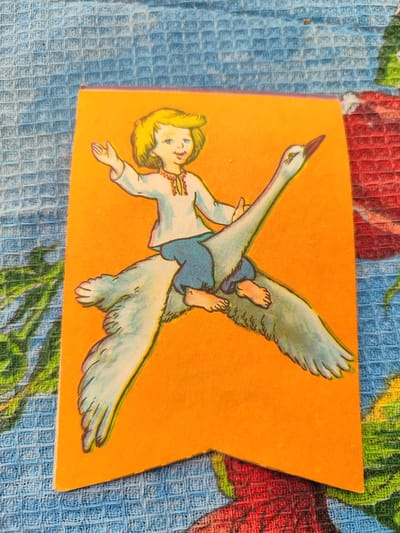 An illustration on an orange banner depicts a blonde-haired child riding a white swan. The child is wearing a white shirt with red and yellow embroidery on the collar and blue pants. The child's left arm is raised. The swan has its wings spread wide, and its head is turned to the left, with its beak pointing upwards. The illustration is set against an orange background. The banner is placed on a blue and white checkered fabric, possibly a tablecloth or a rug. The background fabric is partially covered by a floral pattern in red and green. The image appears to be taken in Pelahiivka, Ukraine.