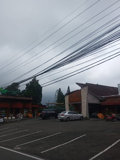 A cloudy sky hangs over a parking lot in front of two buildings. The building on the right, "BUMI AKI", features a wooden facade. Cars and motorcycles are parked in the lot. Power lines dominate the upper portion of the image. An orange umbrella is visible near the "BUMI AKI" building. A person stands near the building on the left.