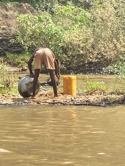 The image depicts an outdoor scene under bright daylight, likely in Kukum, Nigeria. The central subject is an individual, appearing to be an adult with dark skin, wearing light-colored shorts. They are positioned on a dirt or sandy bank next to a body of water.
The person is bent over, seemingly engaged in an activity at the water's edge. They are holding what appears to be a fishing rod or a long stick, suggesting they are fishing or handling fishing equipment. A large, metallic, reflective bowl or pot is situated on the ground near their feet, very close to or partially in the water. Further along the bank, to the left of the person, a rectangular yellow jerrycan or container is visible.
The body of water, likely a river or large pond, is murky and light brown, with reflections on its surface. The bank where the person is standing transitions into dense, green and brown foliage, composed of bushes and trees, which fills the left side of the frame, indicating a natural, perhaps rural or semi-rural, environment. The strong, clear illumination suggests it is daytime, likely sunny. No specific emotions can be discerned, and there is no visible text in the image. The scene captures a moment of practical activity by the water.