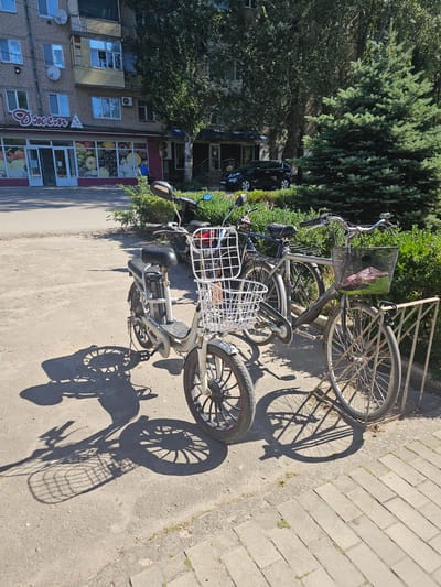 An outdoor, daytime, eye-level shot depicts a row of bicycles and a scooter parked on a paved area in front of a building with a store. The building appears to be a commercial establishment, with large windows displaying fruit and signage that includes the word "Джем" (Dzhem). A black SUV is visible parked on the street behind the bicycles, and a large evergreen tree is prominent in the background. The foreground shows a close-up of the wheels and baskets of the vehicles, with strong shadows cast by the sunlight. The scene is likely located in the city of Skadovsk, Ukraine, given the context provided.