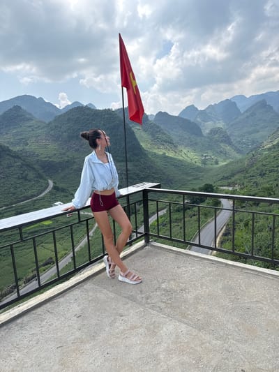 A woman is leaning on a white guardrail, with a mountain range as the backdrop. The location is the Lũng Phìn Commune, Vietnam. She is wearing maroon shorts, a light-colored button-down shirt, and white sandals. A red flag with a yellow star is nearby. A road winds through the green mountainous terrain.