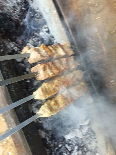 The image captures a close-up, slightly angled view of meat cooking on skewers over hot coals. The scene depicts an outdoor grilling setup, likely a barbecue or mangal.
In the foreground and central part of the image, four metal skewers are visible, each laden with pieces of meat. The meat appears to be cut into roughly rectangular or cylindrical shapes, brownish-grey in color, with some areas showing a slightly darker, more cooked or seared texture. Given the uniform shape, it could be either cubed meat or minced meat (like lula kebab) molded onto the skewers. The skewers are positioned horizontally across a bed of glowing and burnt-out charcoal and ash.
Dense white and grey smoke billows upwards from beneath and around the meat, indicating active grilling and possibly fat dripping onto the hot coals. The coals themselves are a mix of dark black and lighter grey ash, with some spots suggesting residual heat or faint embers, especially on the left side of the frame.
The background on the left consists of more dark, burnt charcoal and ash, suggesting the extent of the grilling area. On the right side, a solid, light-colored surface with a slightly textured appearance is visible, possibly part of the grill structure, a concrete wall, or a wooden surface.
The image is taken from a perspective looking down into the grill, with a noticeable tilt, making the skewers appear to run diagonally across the frame from the upper left to the lower right. No people or visible text are present in the image. The lighting suggests it is daytime, but no specific time or weather conditions can be determined. The overall impression is one of food being prepared over an open flame, with the smoke creating a dynamic and appetizing atmosphere.