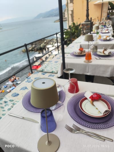 This image depicts an outdoor dining area overlooking the sea in Camogli, Italy. The foreground features a white tablecloth set for a meal with purple glass chargers, white napkins adorned with red flower napkin rings, and silverware. A small, tan lamp with a diffused light sits on the table next to a blue glass. In the background, several tables are set with white tablecloths, suggesting a restaurant or cafe. Beyond the tables, a body of water, likely the Ligurian Sea, is visible, with a rocky shoreline and a beach where several people are sunbathing and swimming. The cityscape of Camogli, characterized by colorful buildings nestled against a hillside, can be seen in the distance. The sky is overcast. The timestamp "31.07.2026 18:55" is visible in the lower right corner. The text "POCO F5" is visible in the lower left corner.