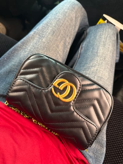 The image shows a close-up of a person's lap, wearing blue denim jeans. A black quilted handbag with a gold double-G logo is resting on the jeans. A bright red fabric is visible beneath the handbag and extending to the bottom left of the frame. The background is dark and out of focus. There is no visible indication of the city of Nookat, Kyrgyzstan in the image.