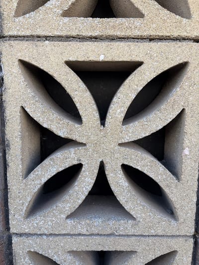 The image shows a close-up view of a stacked concrete block structure, likely a wall or garden feature. The blocks are light grey and have a textured, grainy surface, suggesting they are made of concrete or a similar material. Each block features a decorative cut-out pattern. The central block in the image displays a four-pointed star or flower-like design with curved, leaf-shaped openings. The blocks are stacked horizontally, with visible mortar lines and some minor imperfections such as small cracks and accumulated dust or cobwebs within the openings. The lighting suggests an outdoor environment, possibly under direct sunlight, creating shadows within the recesses of the blocks. There are no people or animals visible in the image. The exact location in Torquay, United Kingdom, cannot be determined from the image alone; it is a detail that would require external information.