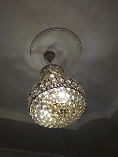 A chandelier is suspended from a ceiling in Rumuomasi, Nigeria. The chandelier has a tiered design with multiple rows of clear, faceted glass beads that reflect the light. The fixture itself appears to be made of a metallic material, possibly brass or gold-toned, with decorative metal trim. The ceiling is a neutral color and features a circular molding around the base of the chandelier. The lighting in the image is dim, creating shadows on the ceiling and surrounding walls. There are no people visible in the image.