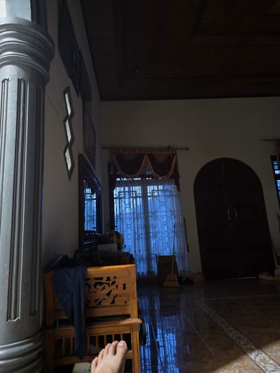 The image depicts an indoor scene with a focus on a wooden furniture item with intricate carvings, likely a cabinet or shelf. A dark blue garment is draped over this furniture. To the left, a large, fluted column is visible, suggesting an architectural element of the room. In the background, there are windows with sheer curtains, and a large, dark wooden door with an arched top. The floor is tiled. A broom leans against the wall near the window. The lighting is dim, casting shadows and creating a subdued atmosphere. The scene is situated within a residence in the city of Bireuenmeunasahblang, Indonesia.
