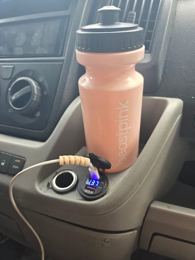 A pink water bottle with a black lid and the text "beastpink" is placed in a cupholder in a vehicle's center console. A white charging cable with beige bead-like protectors is plugged into a car charger, which displays "13.7" in blue LED lights. The charger is located next to a car cigarette lighter port. Buttons labeled "ASR" and a lock symbol are visible to the left of the charging ports. The background shows parts of the car's dashboard, including a vent and a dial. The image is cropped, and the scene appears to be inside a vehicle, possibly a van, during the daytime. The provided location is Straldzha, Bulgaria, but the image itself does not contain any overt visual cues to confirm this specific location; it is a generic interior vehicle shot.