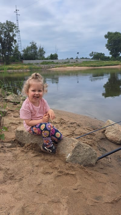 A young girl with blonde curly hair pulled into a high ponytail sits on rocks on a sandy riverbank. She is wearing a pink t-shirt with a floral pattern and colorful leggings with a floral design. Her tongue is sticking out, and she is smiling at the camera. The river is calm and reflects the cloudy sky. In the background, trees and a power line tower are visible. A fishing rod is resting on the sand to the right of the girl. The scene appears to be a park or natural area within the city of Krosno Odrzańskie, Poland, likely near the Oder River.