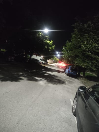 At night, a street scene is captured, likely in the city of Madan, Bulgaria. The asphalt road is illuminated by streetlights and the headlights of parked and passing vehicles. Several cars are visible, including a white van parked on the right side of the road and a blue car further down the street. Trees line the sides of the road, their branches casting shadows on the asphalt. The overall atmosphere is dark, with the artificial lights creating pools of illumination and casting long shadows, indicating a typical urban streetscape at night.
