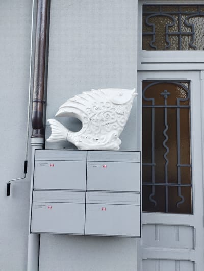A white, decorative fish sculpture sits atop a bank of four grey mailboxes mounted on a textured, pale grey building exterior. The mailboxes are arranged in a two-by-two configuration, each with a horizontal slot and a label. The top left mailbox label reads "Anfidermarti," and the top right label reads "Kremmsi." The bottom left label reads "La Blom." A metal downspout runs vertically along the left side of the building, with a small roller attachment visible near the bottom of the image. To the right of the mailboxes and sculpture is a closed door with a window pane featuring decorative wrought iron bars and amber-colored textured glass. The overall scene appears to be an exterior residential or commercial building entrance. The location is Orselina, Switzerland.