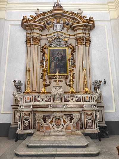 This full-length, slightly tilted indoor photograph captures an ornate Baroque or Rococo-style altar, likely within a church in Monopoli, Italy. The elaborate altar is composed of white and gold painted wooden elements combined with richly inlaid marble (pietra dura or scagliola) panels featuring various colors such as red, brown, black, beige, and green, arranged in intricate scrollwork and geometric patterns.

At the center of the altar is a rectangular painting, set within a gold frame, depicting a religious scene with multiple figures, rendered in somewhat dark tones. Flanking the painting and integrated into the altar's structure are several large, golden candlesticks holding white candles. Numerous sculptural elements, including white and silver-toned putti or angels, adorn the upper and lower sections of the altar, contributing to its grand and opulent appearance. Some angels appear to be supporting elements or decorative motifs.

To the right of the central altar structure, there are architectural elements constructed from light and dark gray marble, including what appears to be steps or a side altar. The altar is set against a light gray wall that features subtle framed paneling. The overall impression is one of rich detail, luxurious materials, and intricate craftsmanship, characteristic of historical ecclesiastical art and architecture found in Italy. There is no discernible activity taking place, and no visible text in the image. The lighting is consistent with an indoor setting, likely diffused or artificial.