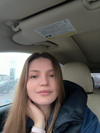 The image displays a young adult female seated inside a vehicle, in what is stated to be the city of Illintsi, Ukraine. No specific landmarks are visible in the image to independently verify this geographic location.
**Subject:**
A female with long, straight, light brown hair and brown eyes is positioned in the foreground, looking directly at the camera with a neutral expression and a slight closed-lip smile. Her left hand is raised, resting with fingers near her jawline/neck. She wears a light pink smartwatch with a metallic face on her left wrist. Her attire includes a dark-colored (possibly black or dark blue) puffy jacket worn over a blue shirt. The subject appears to be in the driver's seat.
**Vehicle Interior:**
The vehicle's interior features light-colored (beige or cream) upholstery on the headliner, sun visor, and the visible portion of the passenger seat headrest. A warning label, affixed to the sun visor above the subject's head, contains multi-lingual text.
**Exterior Environment:**
Through the driver's side window, the view is partially obscured by water droplets on the glass, indicating rain or moisture. Distant, indistinct vertical structures are visible through the window, suggesting an urban or industrial background under an overcast sky.