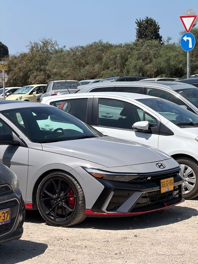 The image captures a sunlit outdoor scene, likely an unpaved parking area or roadside in Kfar Monash, Israel. A row of vehicles is parked under a clear blue sky, with dense green bushes and trees forming a natural backdrop.

In the foreground, a white Hyundai N performance car is prominently featured. It sports distinctive black multi-spoke wheels, red brake calipers, and red aerodynamic trim on its front bumper. The car's front license plate, clearly visible, reads "365-63-004," which is an Israeli license plate format, including the "IL" country identifier. A dark-colored vehicle is parked in front of the Hyundai, with a partially visible Israeli license plate ending in "437." Several other white cars are parked in a line extending into the background, with a yellow car also discernible further down the row.

To the left of the parked cars, two road signs are mounted on a pole: a standard "Give Way" (yield) sign, characterized by its red-bordered white inverted triangle, and a blue circular sign indicating a "Turn Right" with a white arrow. Below these, a yellow sign with Hebrew text is partially visible, reading "ועבר אז ימחה" (v'avar az yimcha), which translates to "and passed then it will be erased/obliterated." The bright sunlight and shadows cast by the vehicles suggest it is midday or early afternoon. No people are visible in the scene.