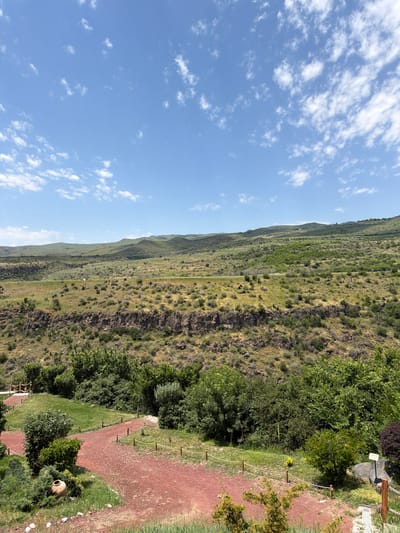 The image captures a scenic landscape in Byurakan, Armenia, featuring rolling green hills under a bright blue sky dotted with white clouds. In the foreground, a reddish-brown path winds through lush greenery and trees. A large, terracotta-colored pot sits on the grass near the path. In the mid-ground, a rocky cliff face rises, covered in sparse vegetation. The background showcases the vast expanse of the Armenian countryside, with layered hills stretching towards the horizon. The scene is peaceful and natural, with no people or animals visible. The lighting suggests it is daytime, likely late morning or early afternoon, given the clear sky and shadows.