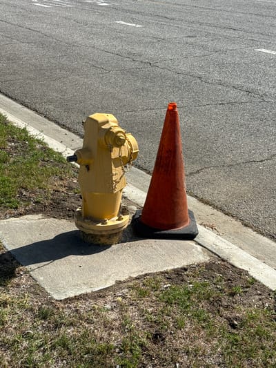 A yellow fire hydrant and an orange traffic cone are positioned on a concrete slab next to a curb. The fire hydrant, which appears to be made of metal, is cylindrical with several caps and a chain attached. The traffic cone, also made of what appears to be plastic, is conical in shape and has a black base. Both objects are situated on a patch of grass and dirt. In the background, a cracked asphalt road is visible, with faint white lines suggesting lane markings. The scene is illuminated by sunlight, casting shadows from the fire hydrant and cone onto the concrete slab. The location is identified as the city of Fontana, United States of America. There are no people or animals visible in the image.