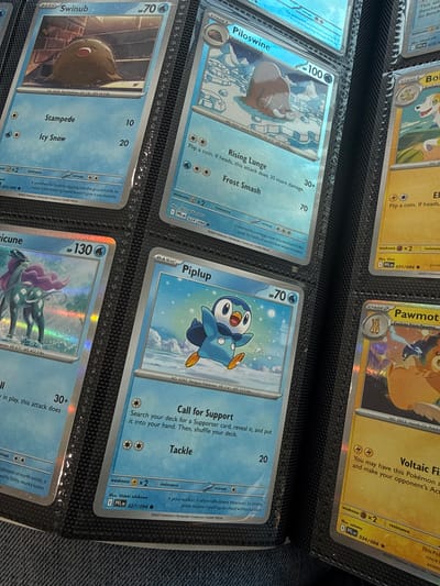 The image shows a close-up view of a black binder filled with Pokémon trading cards. The binder is positioned on a textured surface, possibly a table or floor. Several cards are visible, mostly featuring blue and light blue backgrounds with illustrations of Pokémon.

On the left side, a "SwInub" card is prominent, showing the Pokémon on a brick background. To its right, a "Piloswine" card is visible, depicting the Pokémon in a snowy environment. Further to the right, a yellow card with a dog-like Pokémon is partially seen. Below the Piloswine card, a "Piplup" card is clearly displayed with the Pokémon in a snowy scene. Adjacent to the Piplup card, a yellow card partially shows a Pokémon.

The cards exhibit varying stages (Basic, Stage 1, Stage 2) and information such as HP, attacks, weaknesses, and resistances. The binder's black pages and plastic sleeves are also visible. The lighting suggests an indoor setting. There are no people or any specific landmarks indicating the city of Azzano San Paolo, Italy.