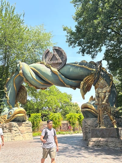 This media file shows an outdoor scene with a statue and a person. The statue appears to be made of bronze and depicts a mythological or fantasy creature with a human-like upper body, possibly a warrior, intertwined with a large, serpentine form. The creature's skin is a teal color, with gold accents. The base of the statue is made of rough, grey stone. In the foreground, a young man wearing a grey t-shirt and khaki shorts stands on a patterned brick walkway. He is looking up and to his right, with a slight smile, suggesting he is observing something or posing for a photo. He has a black backpack slung over his shoulder.

The scene is set outdoors with lush green trees and clear blue sky visible in the background. The lighting suggests it is daytime, likely a sunny day. The presence of trees and the open walkway indicate a park or a public outdoor space. The architectural style of the statue and the surrounding stonework hint at a themed environment, possibly an amusement park or a historical reenactment site. Given the location context of Castelnuovo del Garda, Italy, it could be part of Gardaland or a similar attraction. There is no visible text in the image. The overall mood seems relaxed and pleasant, indicative of a tourist enjoying a day out.