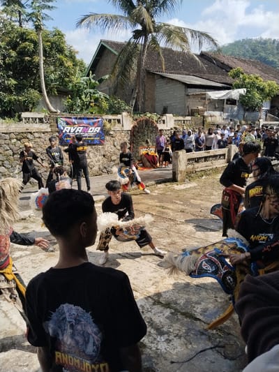 The image captures a traditional Javanese performance, possibly a Jaranan dance, unfolding in what appears to be the village of Jenawi, Indonesia. The performers, some wearing stylized horse costumes, are dressed in black shirts with "ANOM JOYO" printed on them, indicating the name of their troupe. Some participants are positioned in expressive poses, suggesting movement and storytelling. The backdrop features a stone wall decorated with a banner advertising "AVANDA MUSIK, ENTERTAINMENT," possibly a sponsor for the event. A crowd of onlookers is visible beyond the stone wall, adding to the festive atmosphere. Houses and trees are located in the background, typical of a rural Indonesian setting. The ground appears to be a paved or unpaved area in the village.