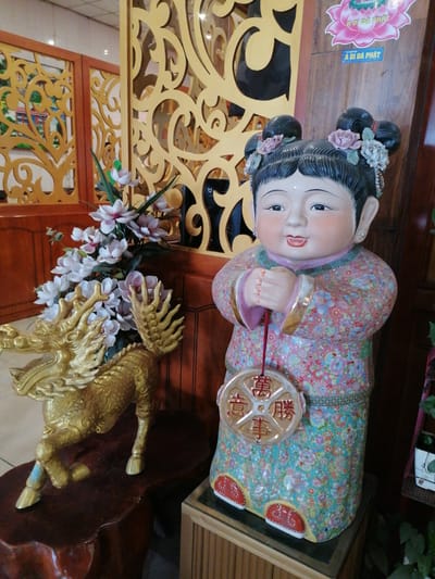The image captures a scene in what appears to be an establishment in Chu Văn An Ward, Vietnam.

To the left, a golden dragon statue sits on a dark, intricately carved wooden stand. Adjacent to the dragon is an arrangement of white and pale pink flowers.

To the right of the dragon, a statue of a child-like figure dressed in a colorful, patterned garment stands on a small wooden pedestal. The figure has two buns with flowers. A round coin hangs from the figure's hands.

The background features wooden decor and a sign at the top with "A Di Đà Phật" on it.